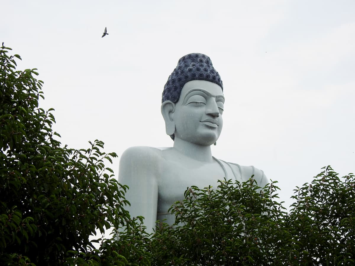 Large white Buddha statue in Phuket, Thailand, rising above tropical trees beneath an overcast sky.