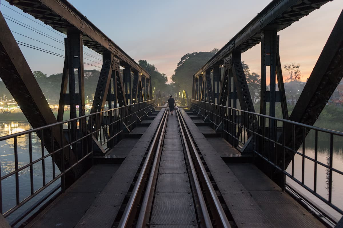 Bridge over the River Kwai in Kanchanaburi, Thailand, with railway tracks crossing the river at sunset.