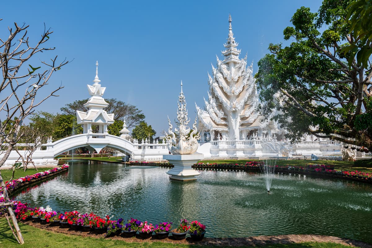 White Temple in Chiang Rai, Thailand, with ornate white architecture, bridge and reflective pond under a clear blue sky.