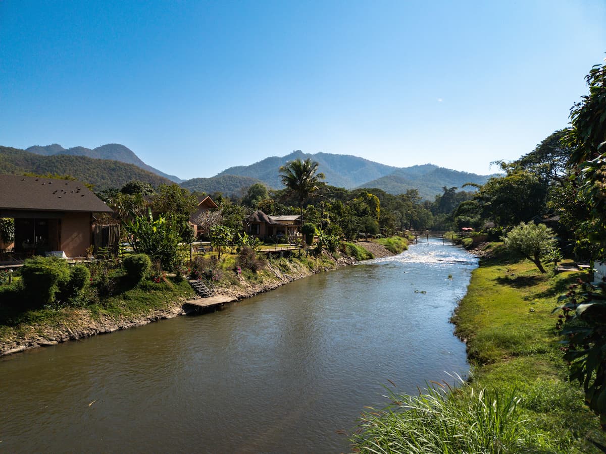 River flowing through Pai in northern Thailand, with riverside accommodation, tropical greenery and mountains under a clear blue sky.