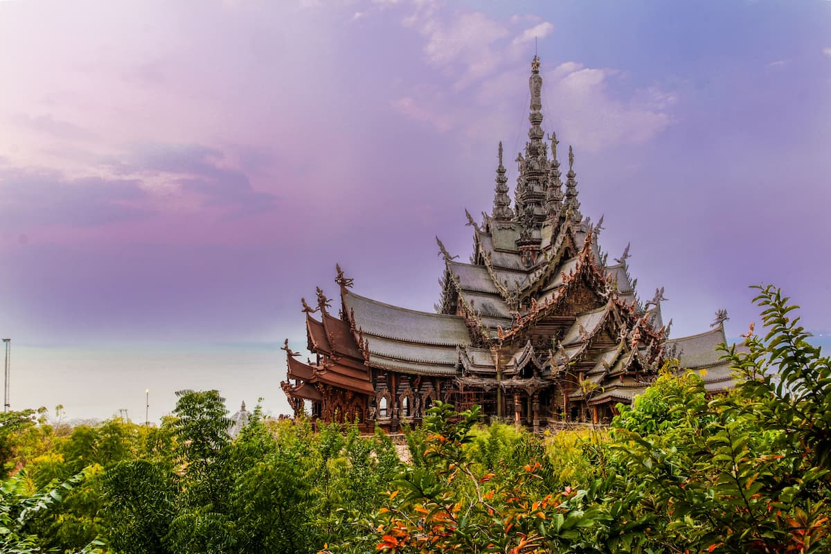 Sanctuary of Truth in Pattaya, Thailand, showing the intricately carved wooden temple surrounded by tropical greenery beneath a pastel sky.