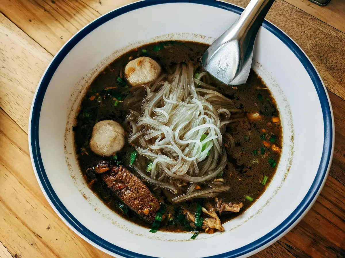 Late-night Thai beef noodle soup with rice noodles and meatballs in a white enamel bowl on a wooden table.