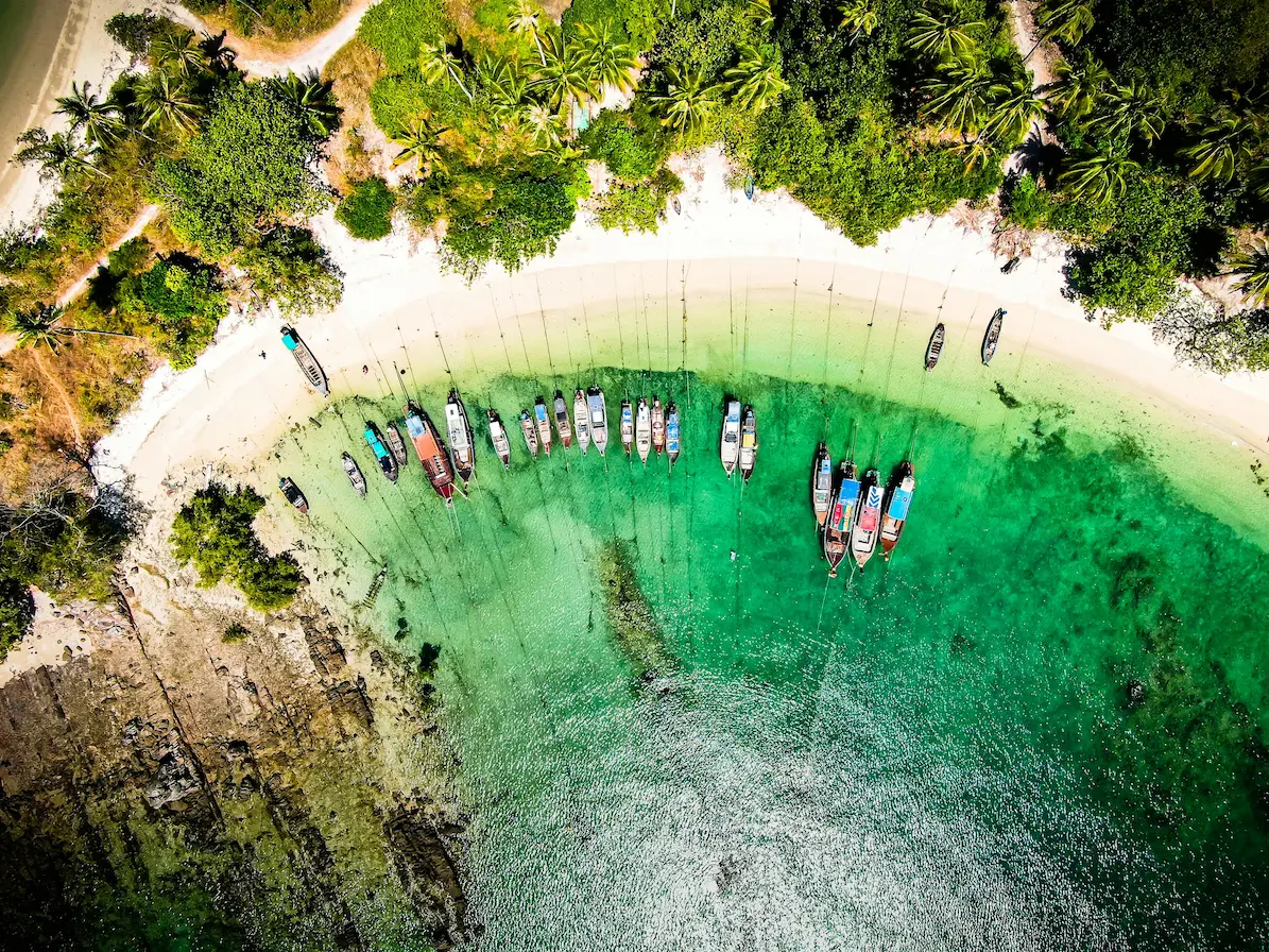 Aerial view of longtail boats lined in a curved emerald bay at Koh Yao Yai, Thailand, surrounded by white sand and palm trees