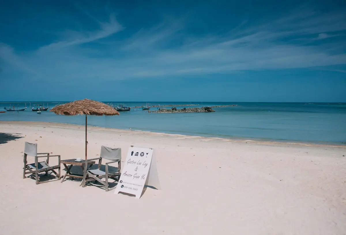 White-sand beach at Koh Samui with two chairs and a straw umbrella facing a calm blue bay and longtail boats