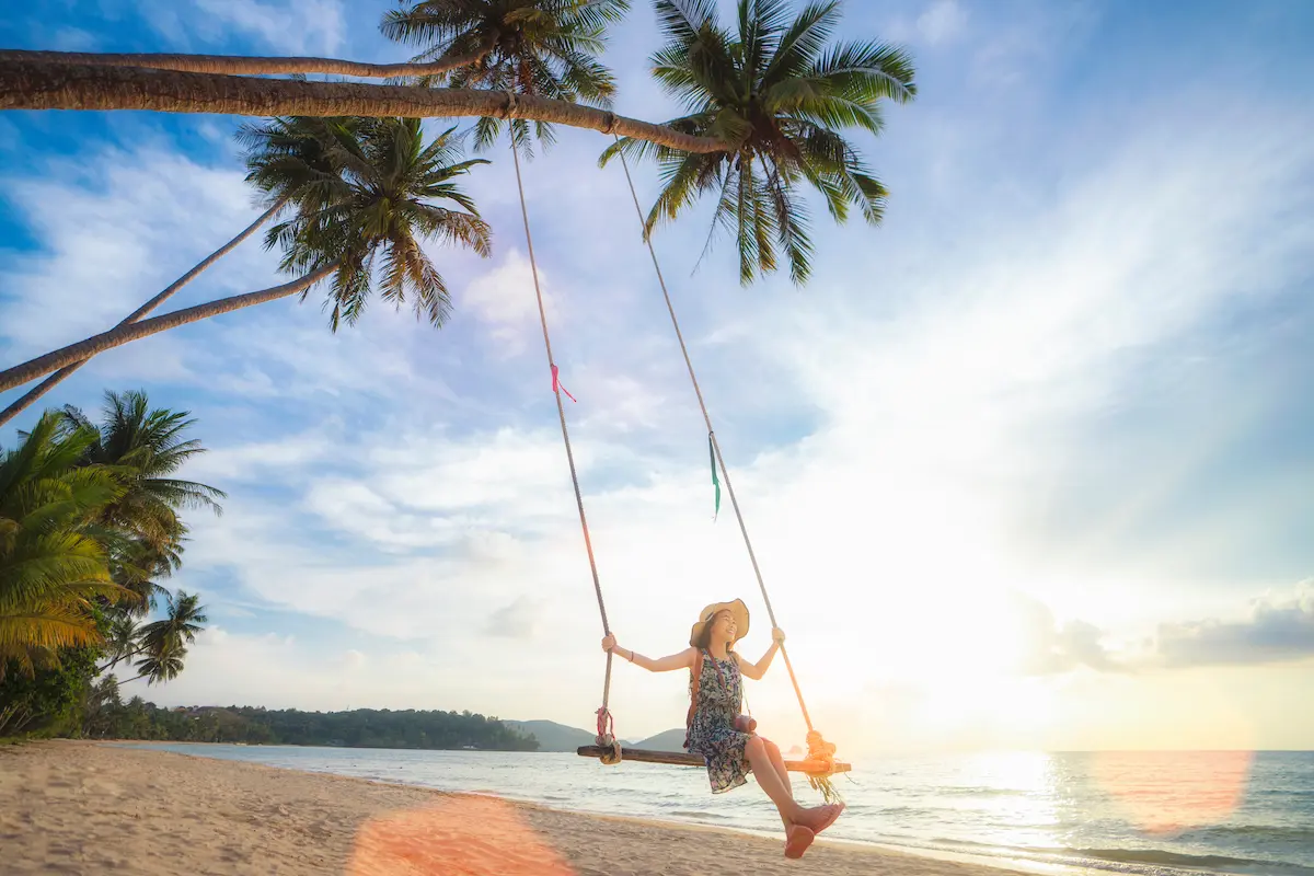 A woman in a sun hat enjoying a swing hanging from palm trees on a tropical beach at sunset on Koh Kood, Thailand.