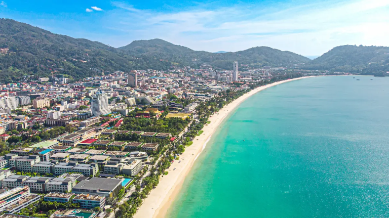 Aerial view of Patong Beach Phuket showing the long sandy shoreline, turquoise bay and city skyline under clear blue sky.