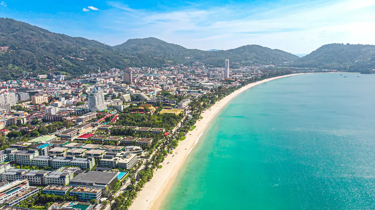 Aerial view of Patong Beach Phuket showing the long sandy shoreline, turquoise bay and city skyline under clear blue sky.