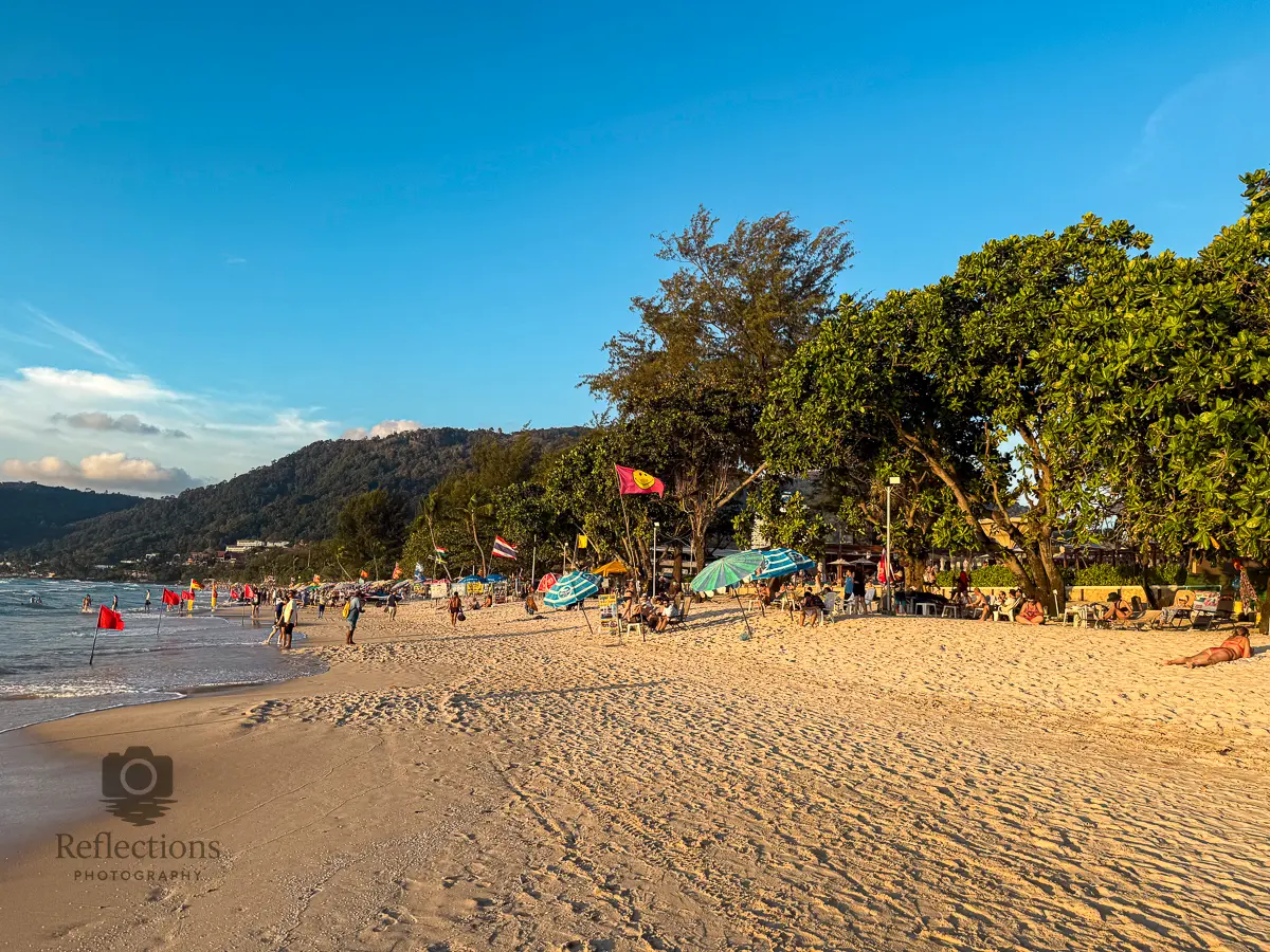 People walking and relaxing on Patong Beach Thailand during golden hour, with warm sunlight, soft waves, and green trees along the shoreline.