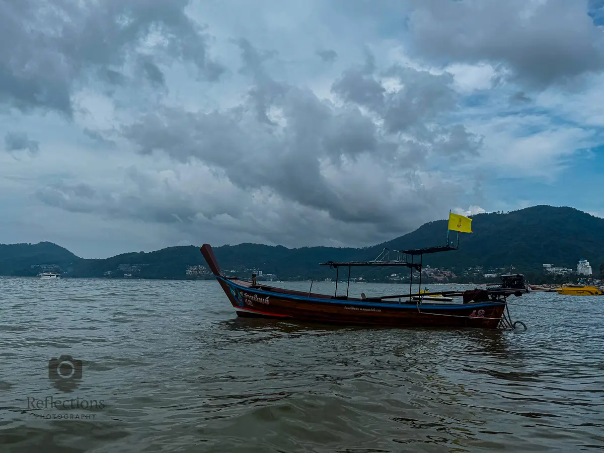 Traditional longtail boat floating on the water at Patong Beach Phuket with mountains and cloudy sky in the background.