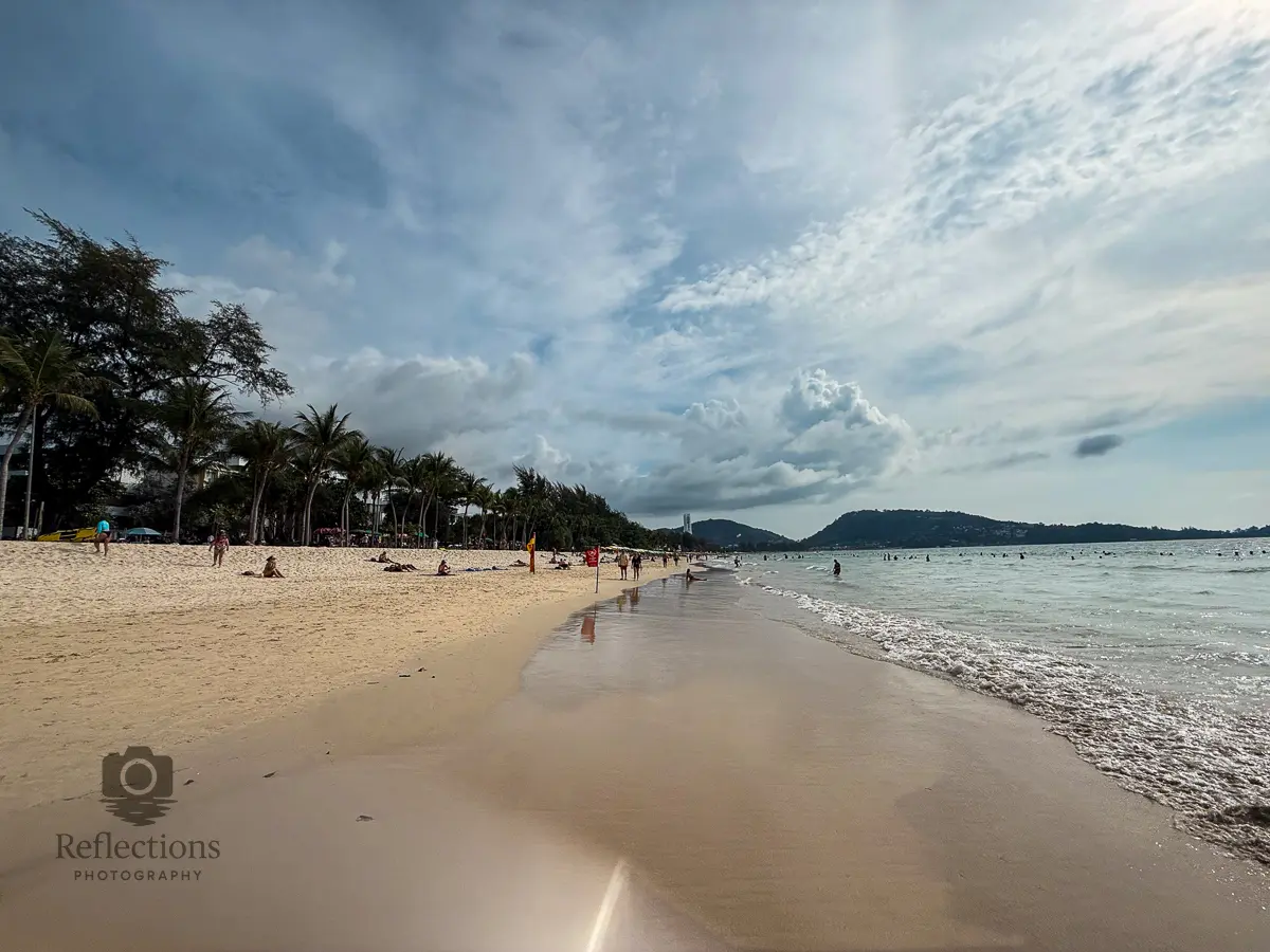 Wide shoreline view of Patong Beach Thailand with soft waves, palm trees and swimmers under a dramatic cloudy sky