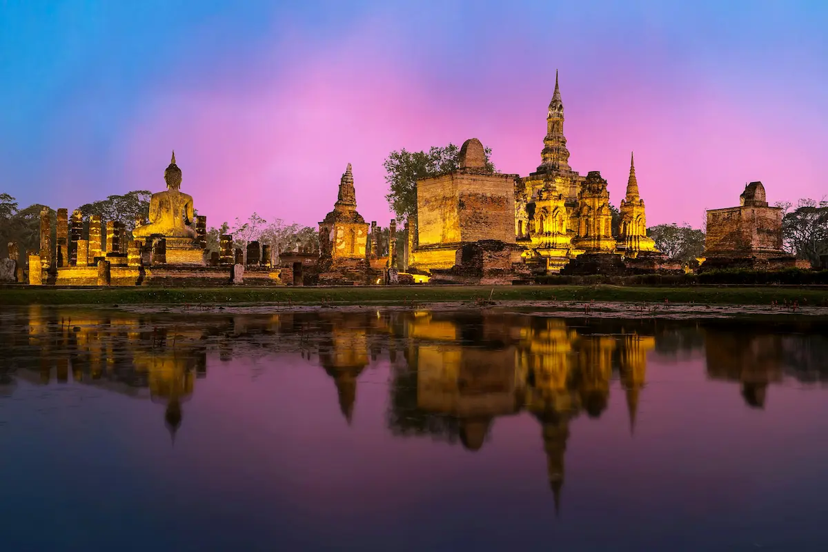 Ancient temples of Sukhothai Historical Park in Thailand reflecting in a lake at sunset.