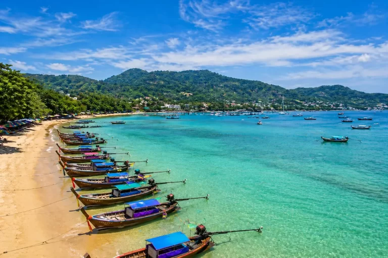 Longtail boats lined along the shore at Rawai Beach Phuket with turquoise water and green hills in the background
