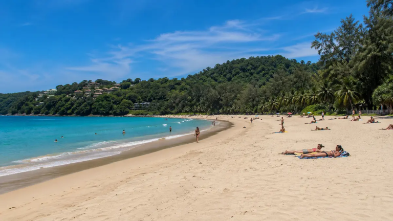 Surin Beach Phuket with clean sandy shoreline, turquoise water, tropical hills, and travellers relaxing during daytime