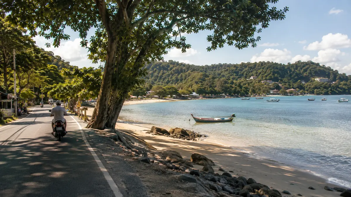 Coastal road and calm bay in South Phuket near Rawai and Nai Harn, with longtail boats, tropical hills and a quiet local beach atmosphere.
