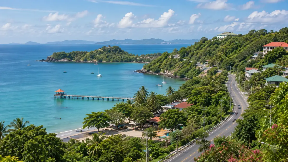 Coastal view of South Phuket near Cape Panwa and Ao Yon, with calm blue water, green hills, boats, a pier and a quiet seaside road.